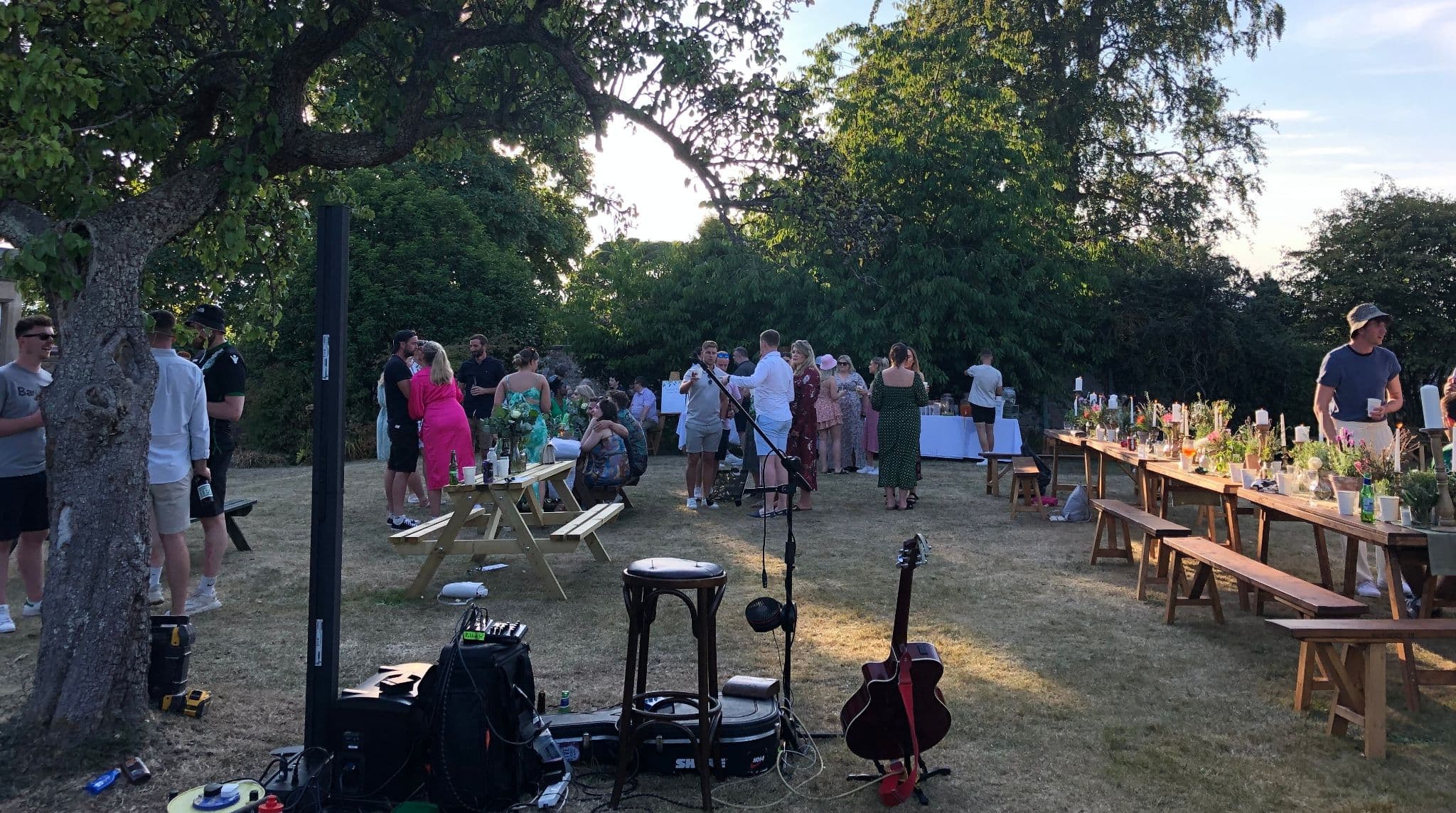 Outdoor wedding drinks reception under a tree with long trestle tables and live music set up
