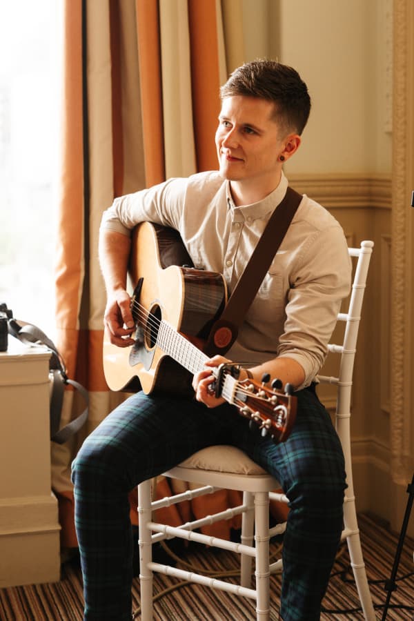 Robbie Wisniewski seated by a window playing acoustic guitar at a wedding venue