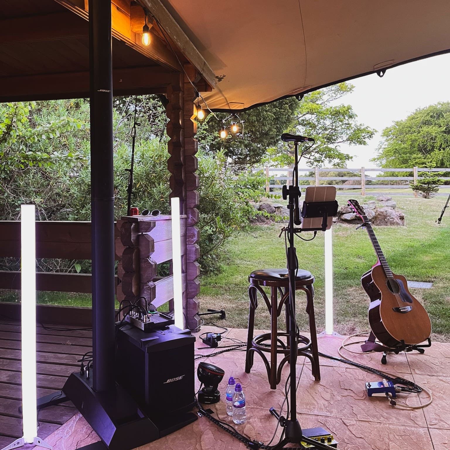 Robbie Wisniewski’s live setup on a covered outdoor deck: PA speaker, LED uplighters, stool, mic stand and acoustic guitar